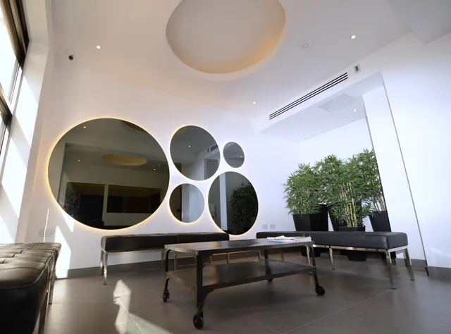 A modern lobby seating area with circular backlit mirrors on the wall, black leather benches, a metal coffee table and potted bamboo plants.