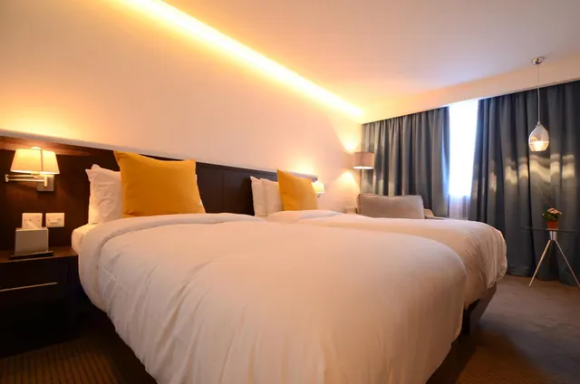 A hotel bedroom with two beds side by side, yellow cushions, wall lamps, grey curtains and a small upholstered armchair in the background.