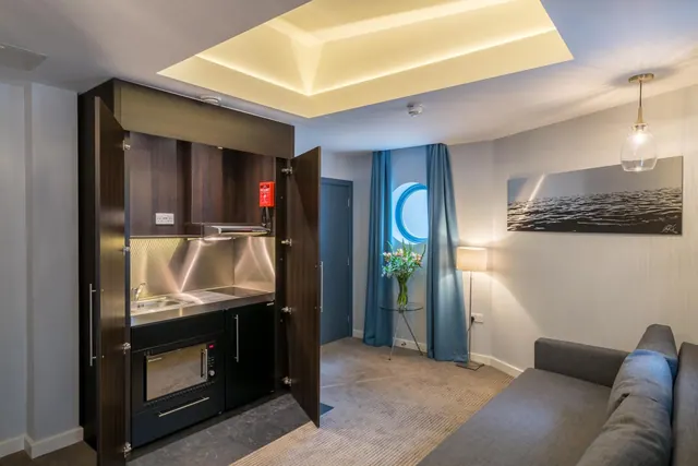 A room living area with an open wooden cabinet holding a sink, hob, microwave and fridge, a grey sofa, a round porthole window with blue curtains, a floor lamp and a framed seascape picture.