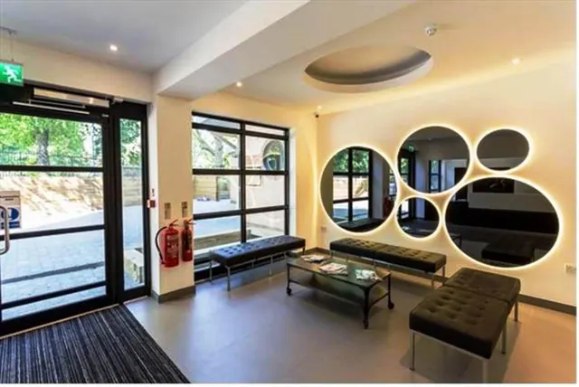A lobby waiting area with black tufted benches, a glass coffee table with magazines, backlit circular mirrors on the wall, and glass doors and windows looking out onto trees and a paved courtyard.