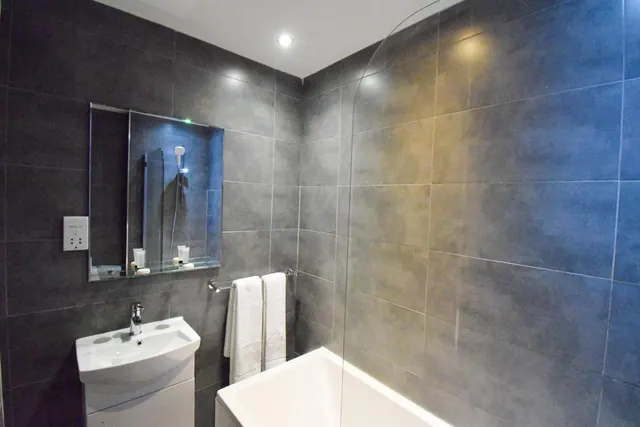 A bathroom with dark grey tiled walls, a white wash basin with a wall mirror and shelf, a white bath with a glass screen, and white towels hanging on a rail.