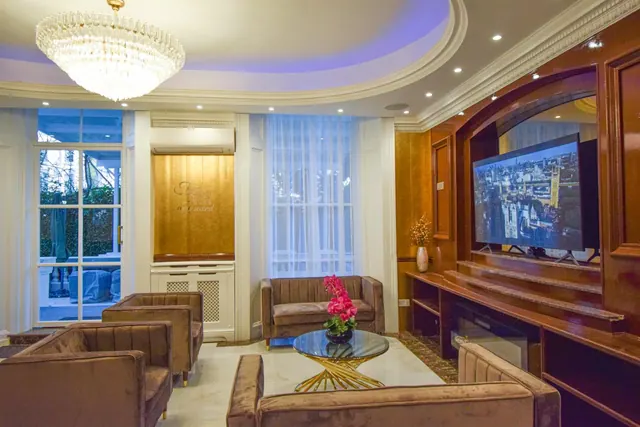 A lounge seating area with velvet armchairs, a sofa, a glass coffee table, a wall-mounted television and a chandelier, with tall windows and French doors looking onto greenery.