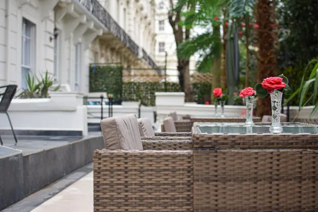 Wicker chairs and a glass-topped table with red roses in vases sit on a paved terrace beside a white stucco building.