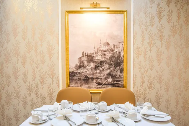 A dining table set with white cups, saucers, folded napkins and glasses sits in front of a framed sepia print of a historic waterfront city, with gold-patterned wallpaper on the walls.