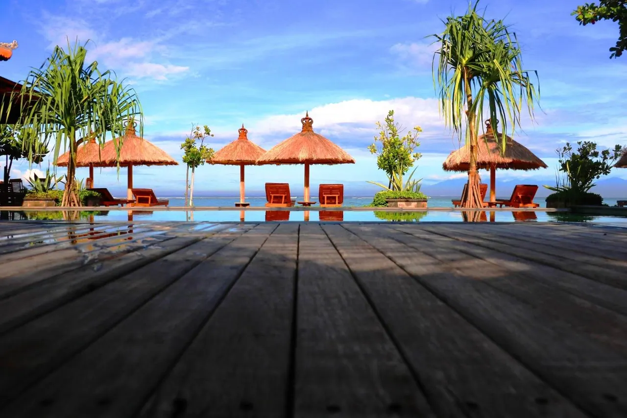 A pool with thatched umbrellas and sun loungers along its edge, with the sea and distant hills beyond, viewed from a wooden deck.