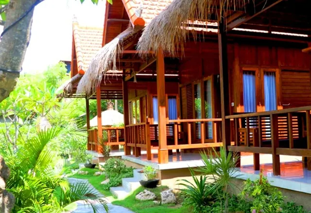 A wooden Balinese-style cottage with a thatched roof, a raised veranda with railings, and blue curtained doors, set among tropical plants beside a stone path.