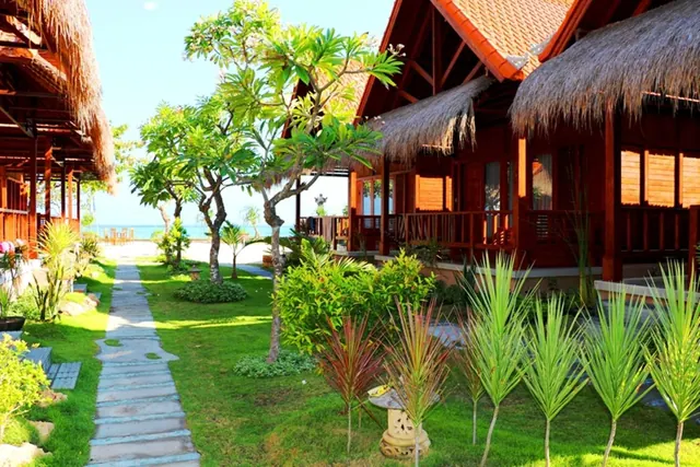 A stone path runs through a lawn between thatched-roof wooden bungalows, with frangipani trees and the sea visible in the distance.