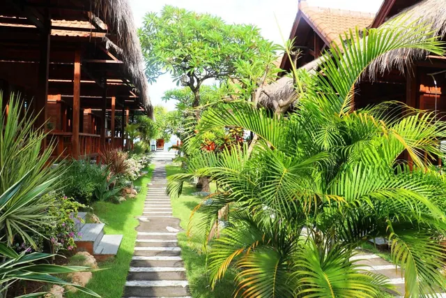 A stone stepping path runs between trimmed grass and tropical plants toward thatched-roof wooden bungalows.