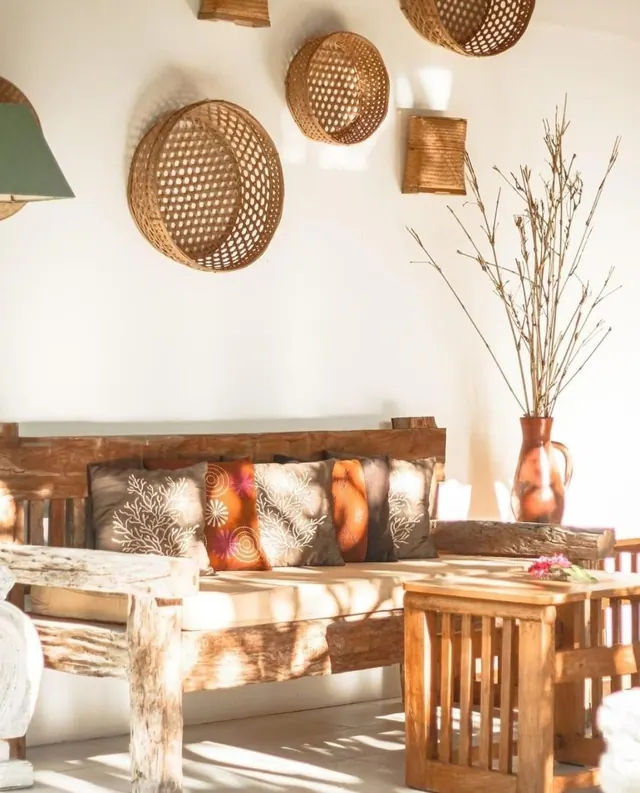 A wooden bench sofa with patterned cushions sits beside a wooden side table, with woven baskets on the white wall and a vase of dried branches in the corner.