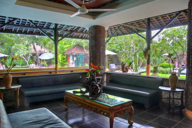 A seating area with dark leather sofas and a carved wooden coffee table, with a ceiling fan overhead and a garden and pool visible through the windows.