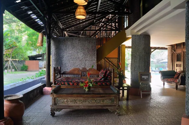 An open-air lounge with a carved wooden daybed and a low table holding a flower arrangement, with a staircase and garden visible beyond.