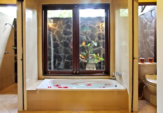 A bathroom with a built-in bathtub containing red flower petals, a large window onto a stone wall and potted plant, a walk-in shower on the left, and a toilet with bidet seat on the right.