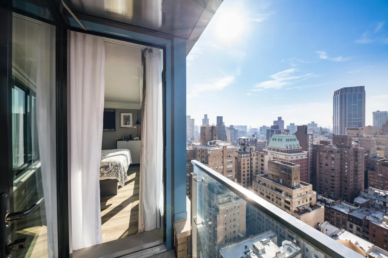 A glass-railed balcony overlooks dense city buildings under a bright sky, with an open door leading into a bedroom with a bed and white curtains.