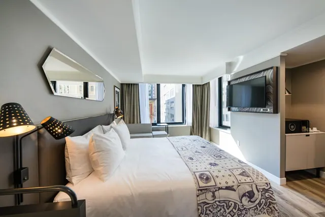 A made king bed with white pillows and a patterned throw faces a grey sofa, a wall-mounted television and a window looking onto city buildings.