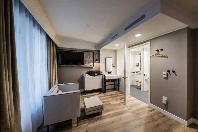 A hotel room with a grey armchair, ottoman, wall-mounted television and white cabinet, with an open doorway to a bathroom containing a walk-in shower and grab rails.