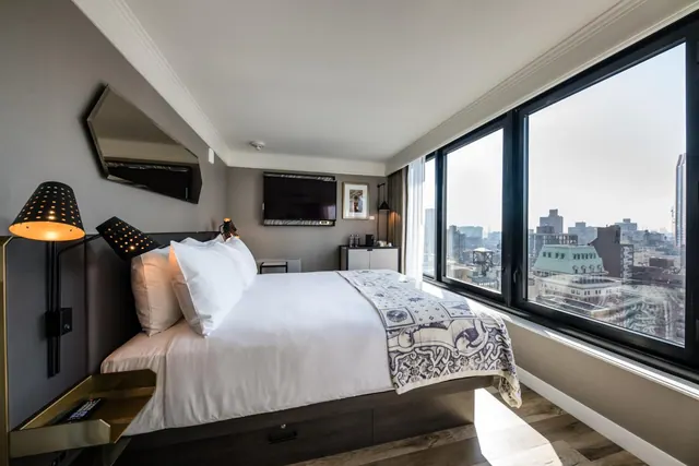 A bedroom with a made king bed, a wall-mounted television, and floor-to-ceiling windows looking out over a city skyline.
