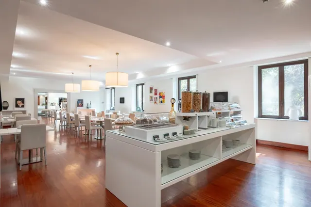 A bright breakfast room with white tables and chairs, a white buffet counter with cereal dispensers and stacked plates, and tall windows showing greenery outside.