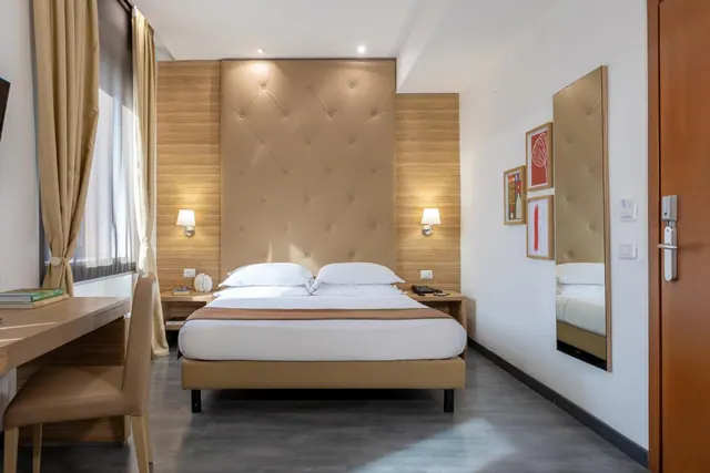A double bed with a tufted headboard sits between two bedside lamps, with a desk and chair on the left and a wall mirror on the right.