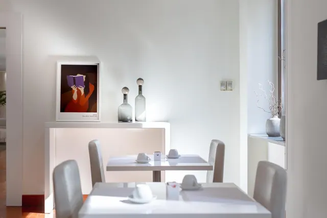 A white dining room with grey upholstered chairs, two small tables set with cups and saucers, a framed print and glass bottles on a white console, and a vase of dried branches by the window.