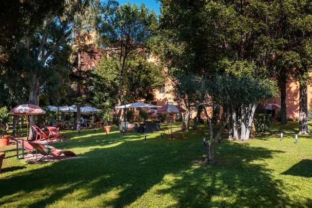 A grassy garden with red-striped sun loungers, parasol umbrellas, tall trees and a terracotta-coloured building behind, with a few people seated in the distance.