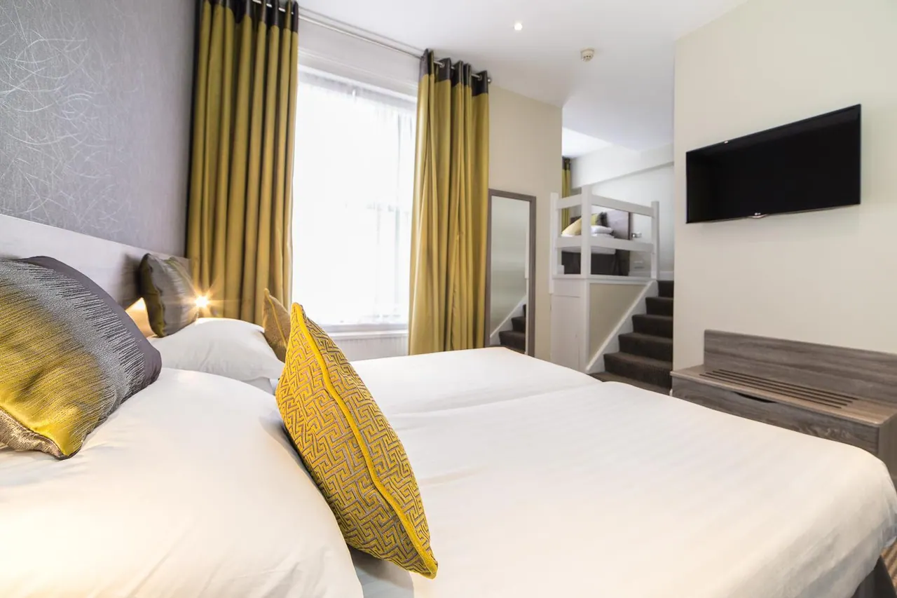A bedroom with two beds made with white bedding and yellow patterned cushions, a wall-mounted television, gold curtains at the window, and a staircase leading up to a raised sleeping area.