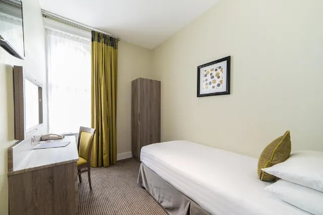 A single bed, wooden wardrobe, desk with a chair and a wall-mounted television in a small bedroom with a window and green curtains.