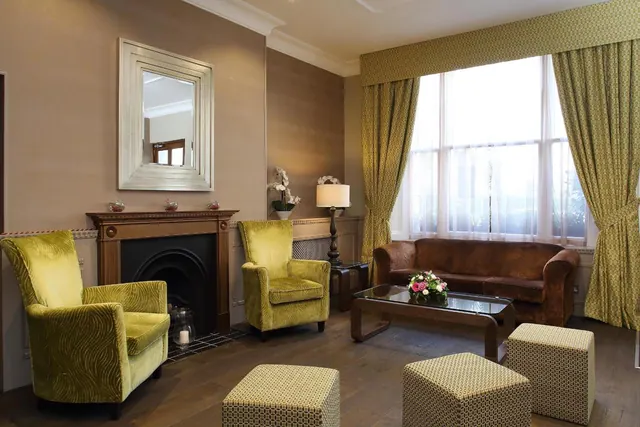 A lounge area with two yellow velvet armchairs, a fireplace, a brown leather sofa, a glass coffee table and a bay window with sheer curtains.