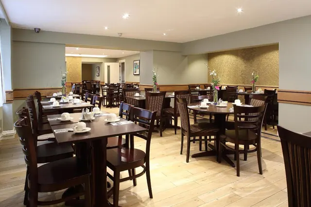 A dining room with rows of dark wooden tables and chairs, set with cups and saucers, on a light wood floor.