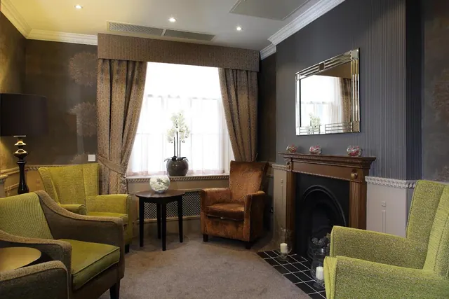 A lounge seating area with green and brown armchairs, a black-tiled fireplace, a side table, and a window with cream sheers and patterned curtains.