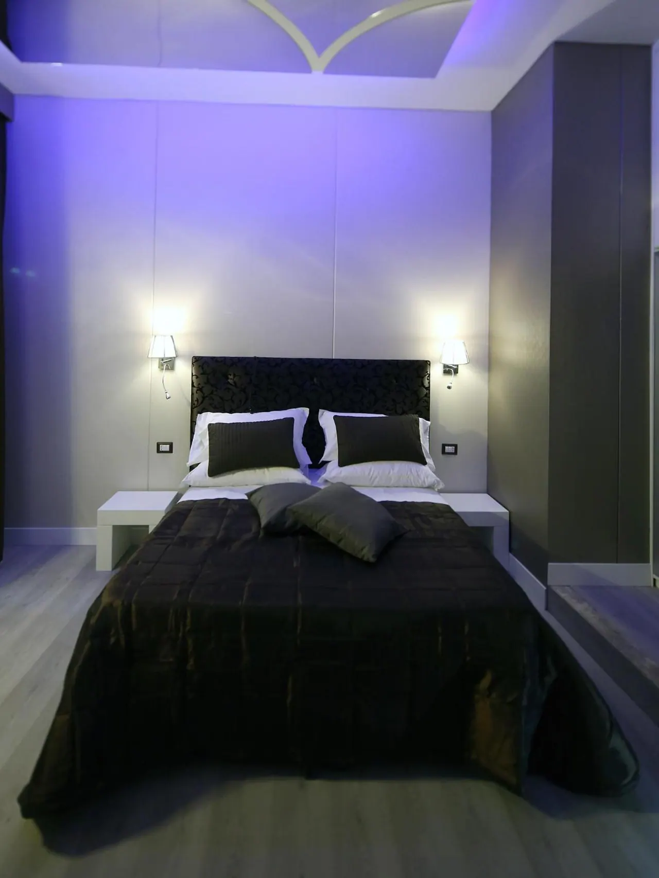 A double bed with a dark quilted cover and black tufted headboard sits in a bedroom lit by purple lights, with wall lamps on either side and no window visible.