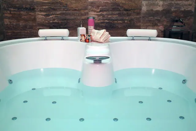 A double whirlpool bath with jets and two headrests, with a soap bottle, pink jar and folded towel on the rim against a brown stone wall.