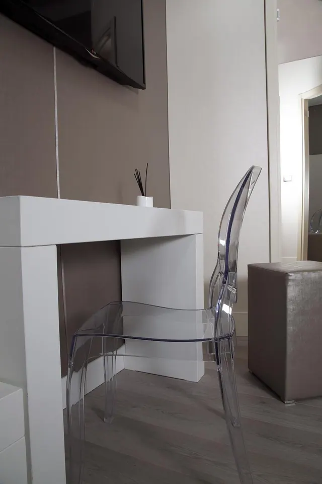 A white desk with a reed diffuser sits beneath a wall-mounted television, with a transparent acrylic chair in front and a taupe ottoman to the right.