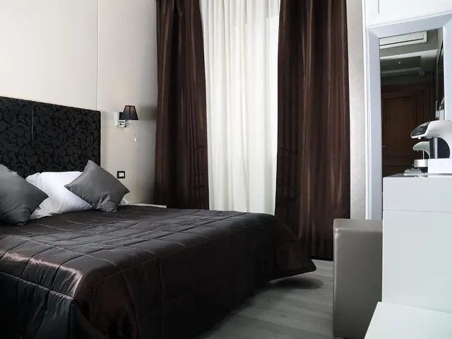 A bedroom with a dark quilted bedspread, a black patterned headboard, grey and white pillows, brown striped curtains and a white coffee machine on a cabinet.