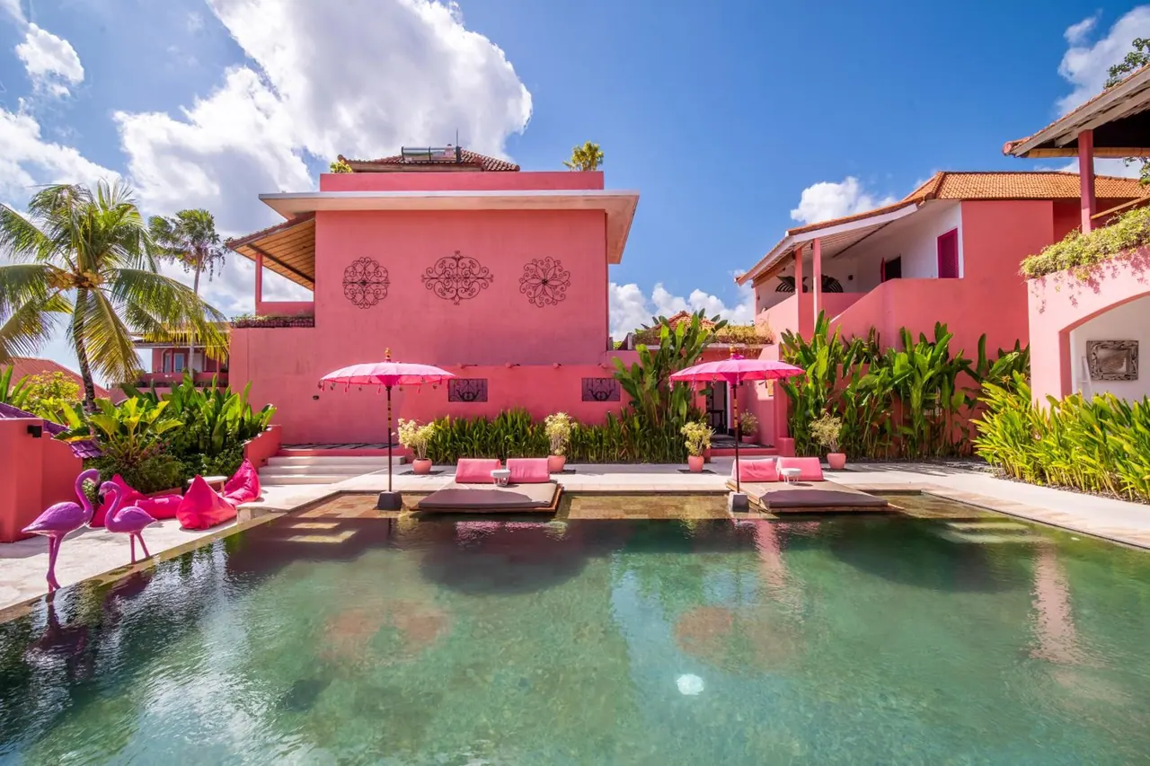 A pool with pink flamingo ornaments, pink umbrellas and cushioned daybeds beside pink-walled buildings under a blue sky.