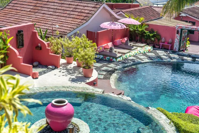 A curved pool with stone edging sits beside pink-walled seating areas and a pink umbrella, with a large pink urn standing in the water at the near edge.