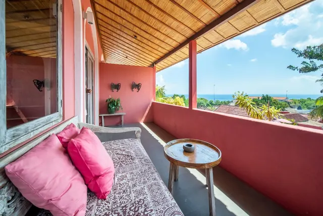 A covered balcony with a patterned daybed, pink cushions and a round wooden side table, looking out over rooftops and palm trees toward the sea.