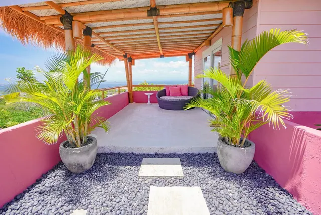 A covered terrace with a bamboo roof, pink walls, potted palms, a round outdoor daybed and a view of the sea beyond the railing.