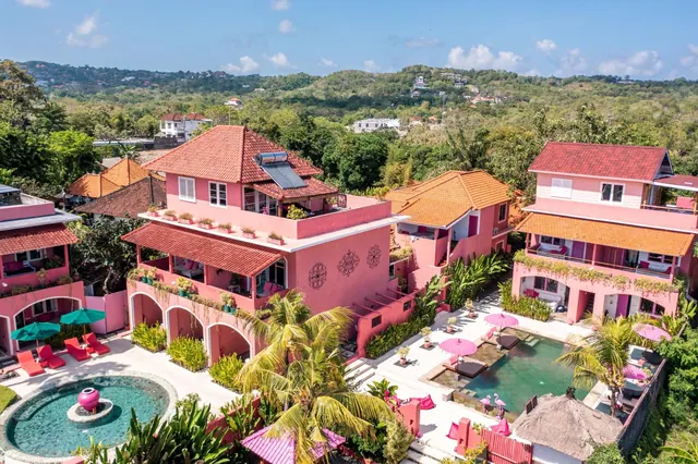 Aerial view of a pink hotel complex with terracotta roofs, a small pool with pink umbrellas and sun loungers, and palm trees, with forested hills behind.
