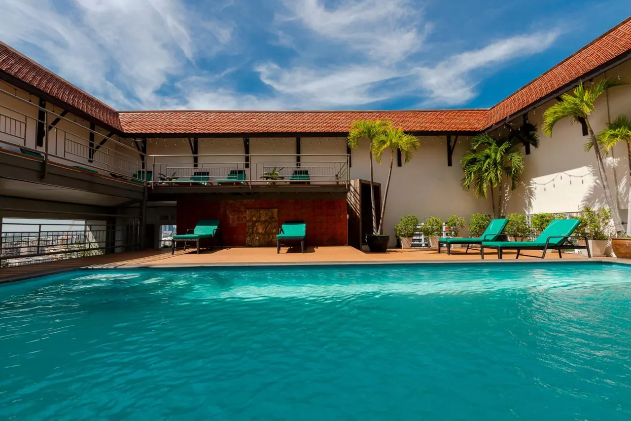 An outdoor pool with teal water, green sun loungers on a wooden deck, potted palms, and a terracotta-tiled roofline under a partly cloudy blue sky.