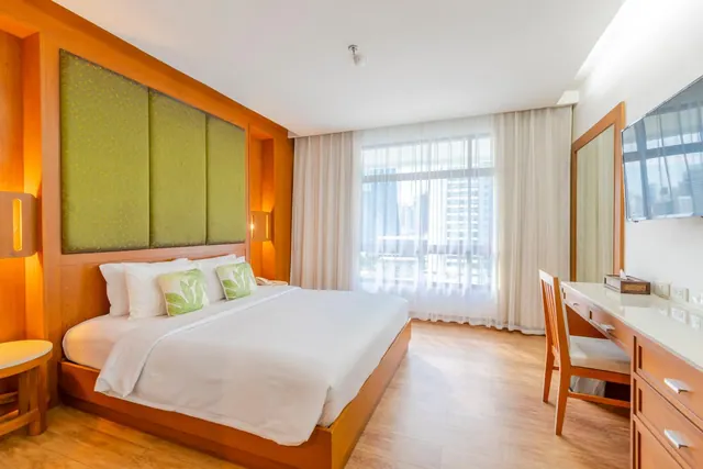 A bedroom with a made king bed, green padded headboard panels, a wall-mounted television, a wooden writing desk and a large window looking out over city buildings.