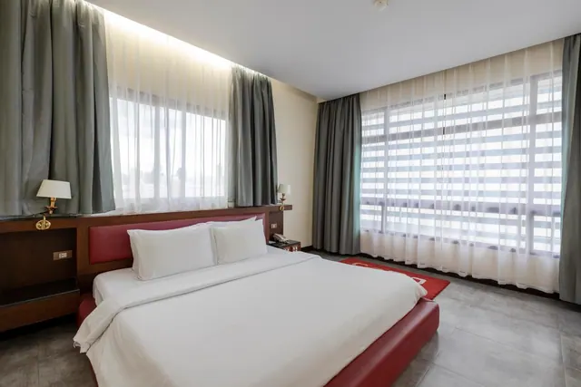 A bedroom with a king bed that has a red leather headboard and base, two bedside lamps, and a large window with sheer curtains and blinds.