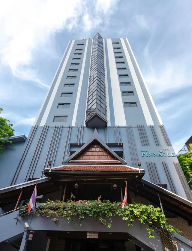 Low-angle view of a tall hotel tower with a Thai-style entrance canopy, flowering plants and flags, under a partly cloudy sky.