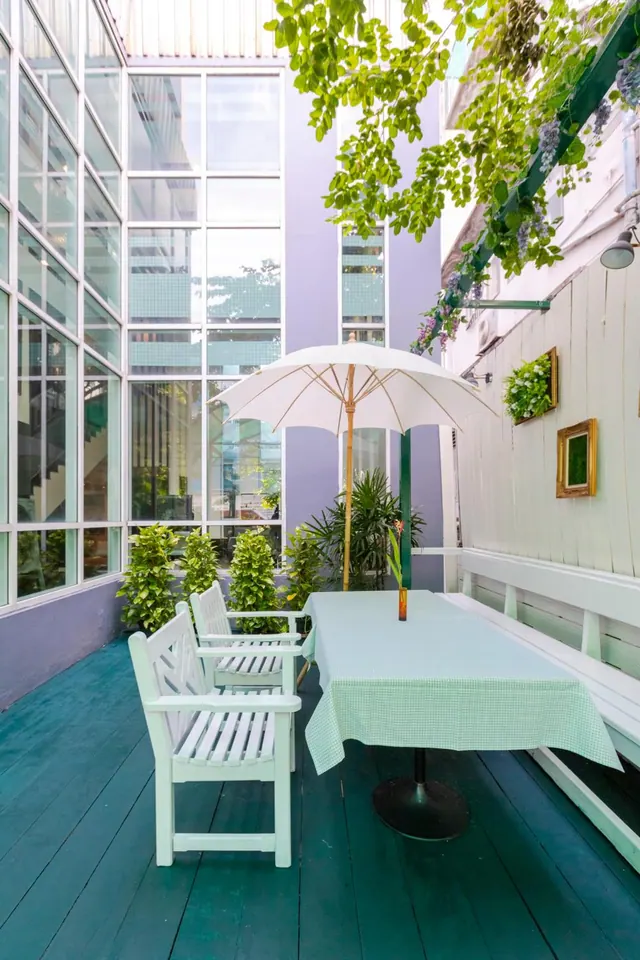 An outdoor terrace with a painted green deck, white benches and chairs, a table with a mint gingham cloth and a white parasol, beside a glass-walled building.
