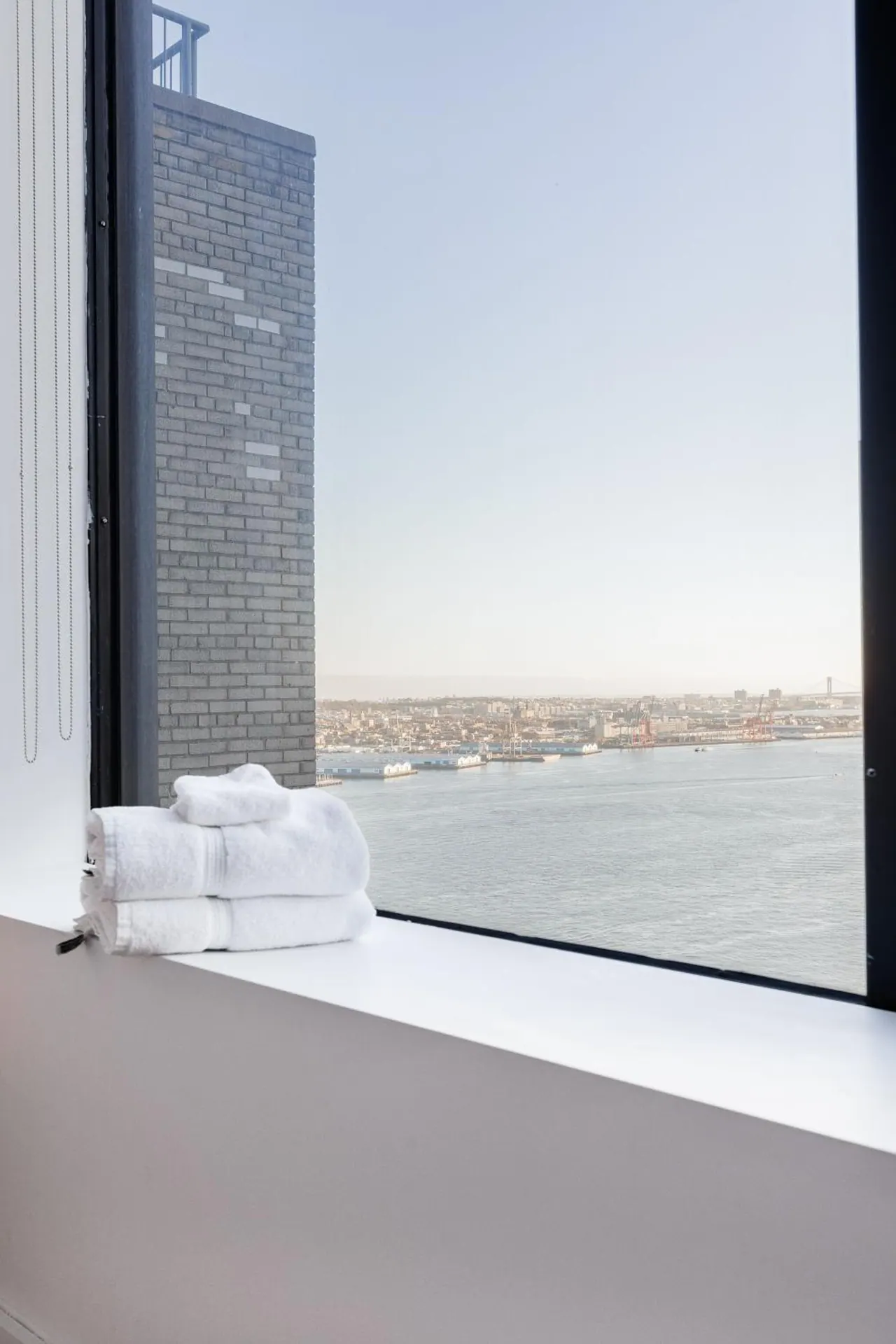 Folded white towels sit on a window ledge beside a tall window overlooking a river, a brick tower and a waterfront city.