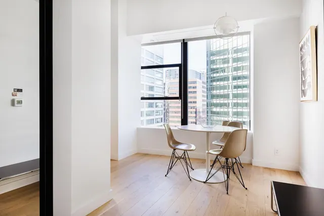 A small dining area with a round white table, four shell chairs and a pendant light beside a large window showing city buildings.