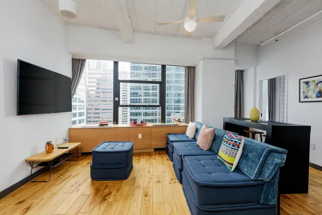 A living area with a blue sectional sofa, an ottoman, a wall-mounted television and a large window looking onto high-rise office buildings.