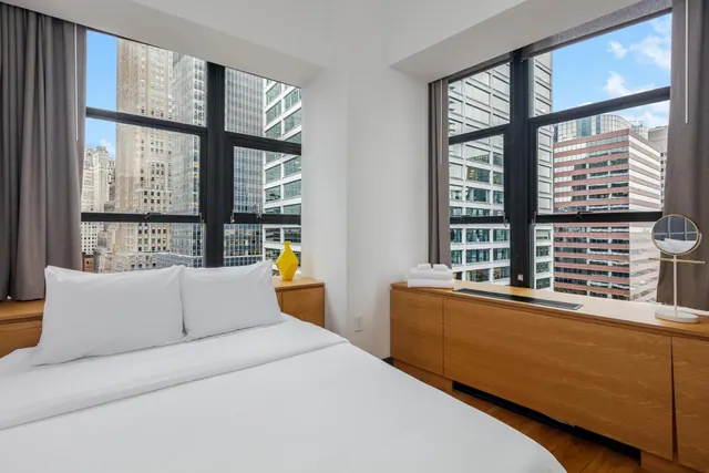 A double bed with white linens sits in a bedroom beside two windows overlooking high-rise buildings, with a wooden cabinet and a round mirror on the right.