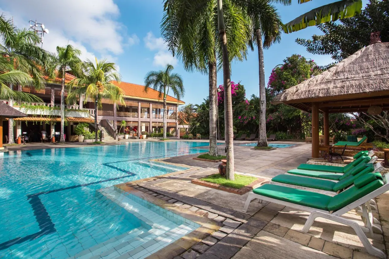 An outdoor pool with palm trees, a thatched gazebo and green-cushioned sun loungers beside it, with a two-storey building behind.