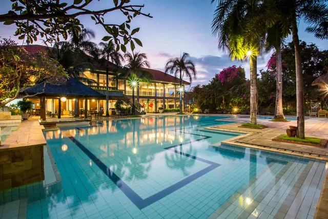 A large tiled swimming pool at dusk with illuminated palm trees and a two-storey building with a pavilion-style roof behind it.