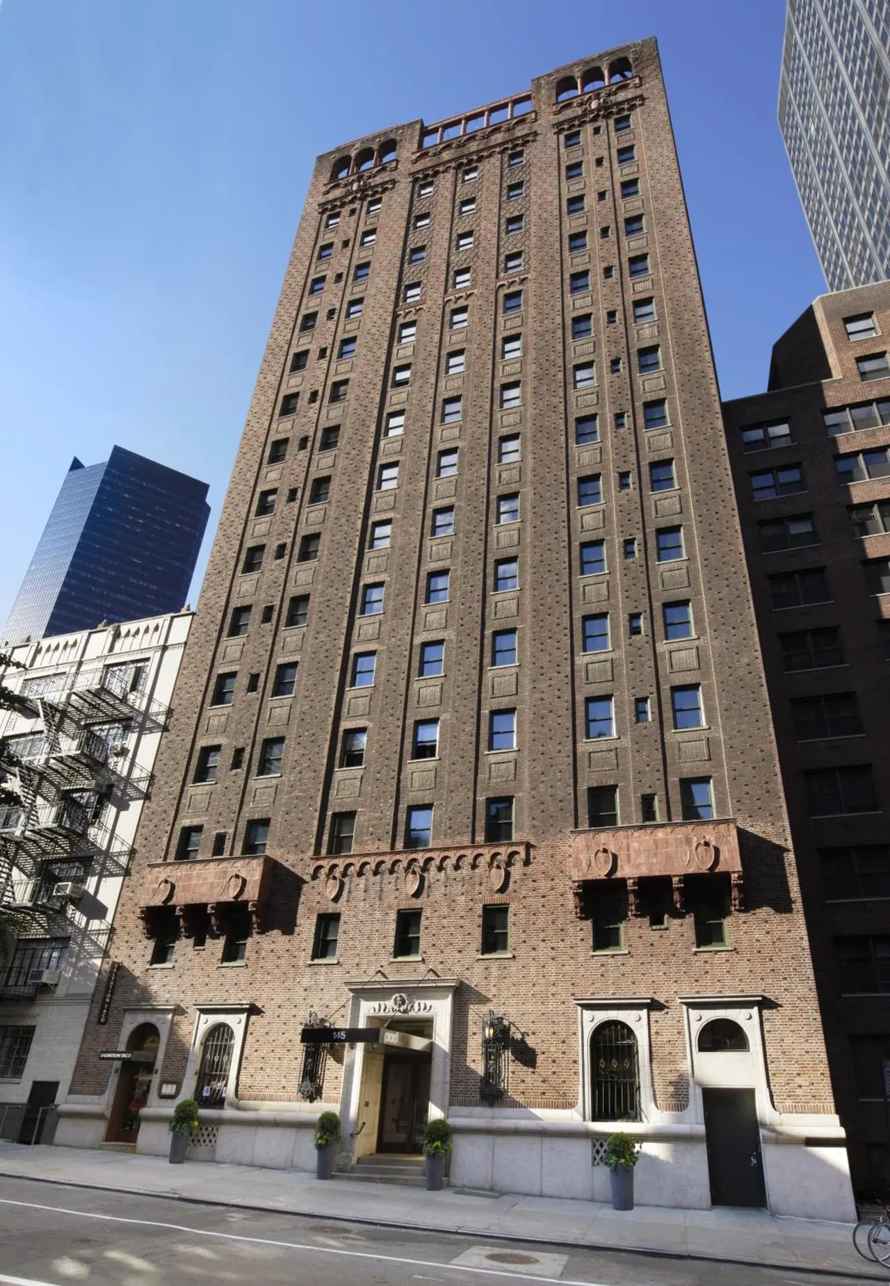 Exterior view of a tall brown brick building with arched entrance doorways and potted plants at street level under a clear blue sky.
